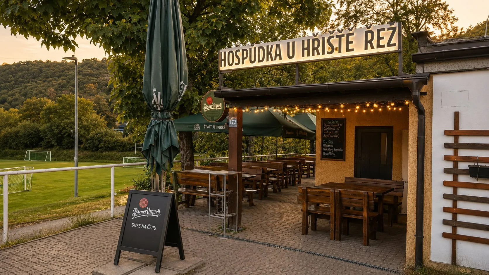 Das Gasthaus von außen — das beleuchtete Schild, überdachte Sitzplätze und daneben der Fußballplatz