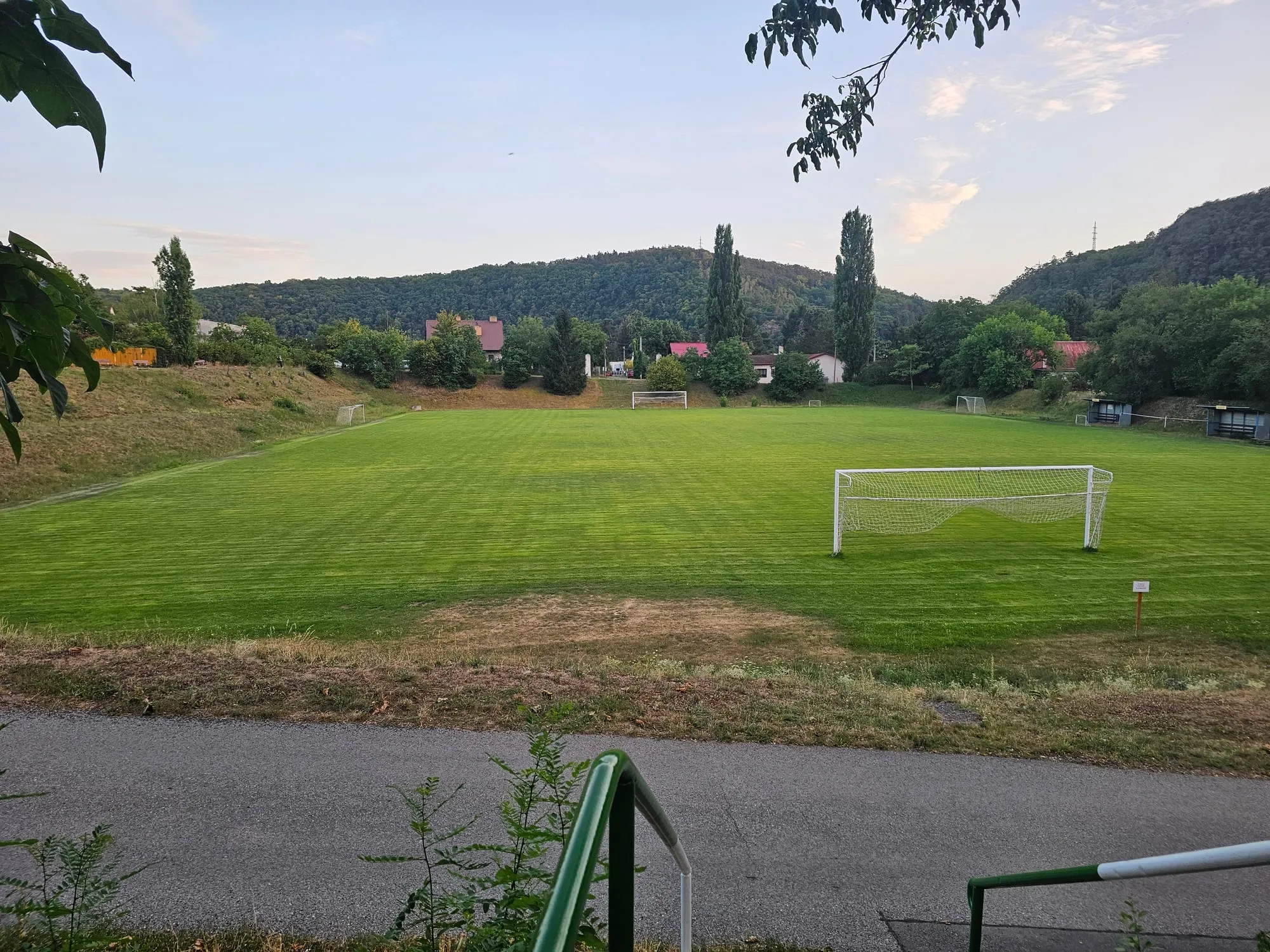 Fotbalové hřiště s brankou, za ním domky Řeže a zalesněné kopce ve večerním světle