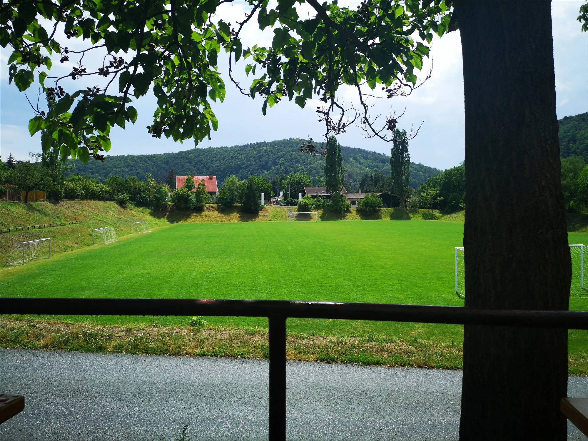 Blick von der Terrasse über das Geländer auf den Sportplatz, dahinter Řež und bewaldete Hügel
